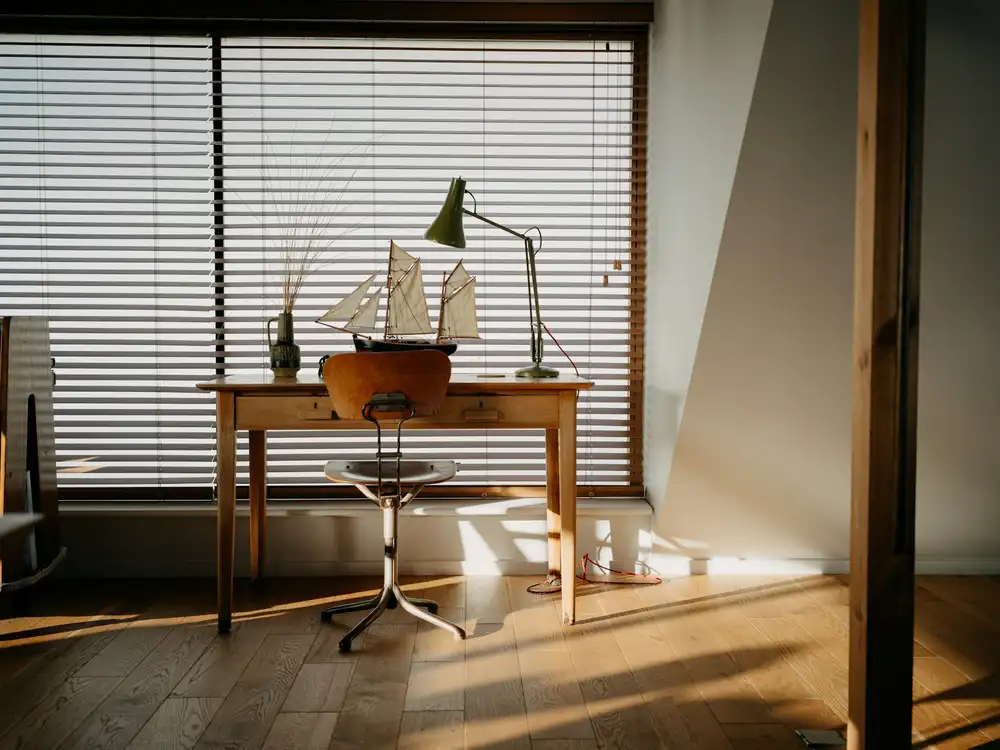 Bureau en bois massif devant une fenêtre à stores, lampe d’architecte et soleil rasant