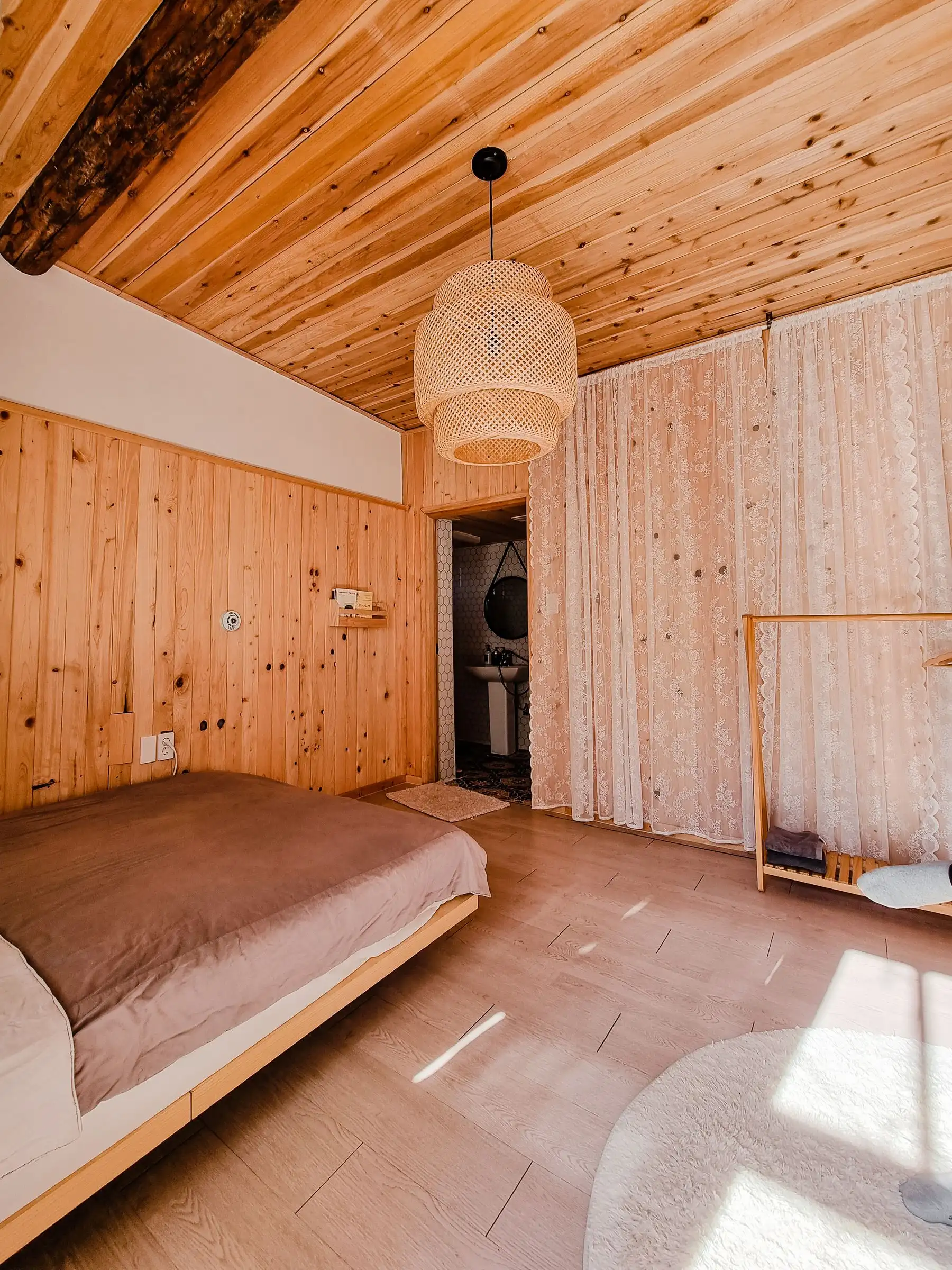 Chambre entièrement habillée de bois clair noueux, poutre apparente et suspension en rotin
