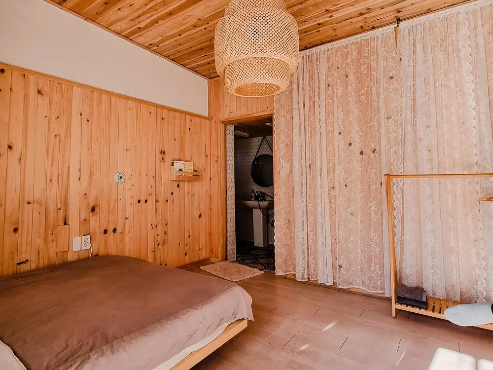 Chambre entièrement habillée de bois clair noueux, poutre apparente et suspension en rotin