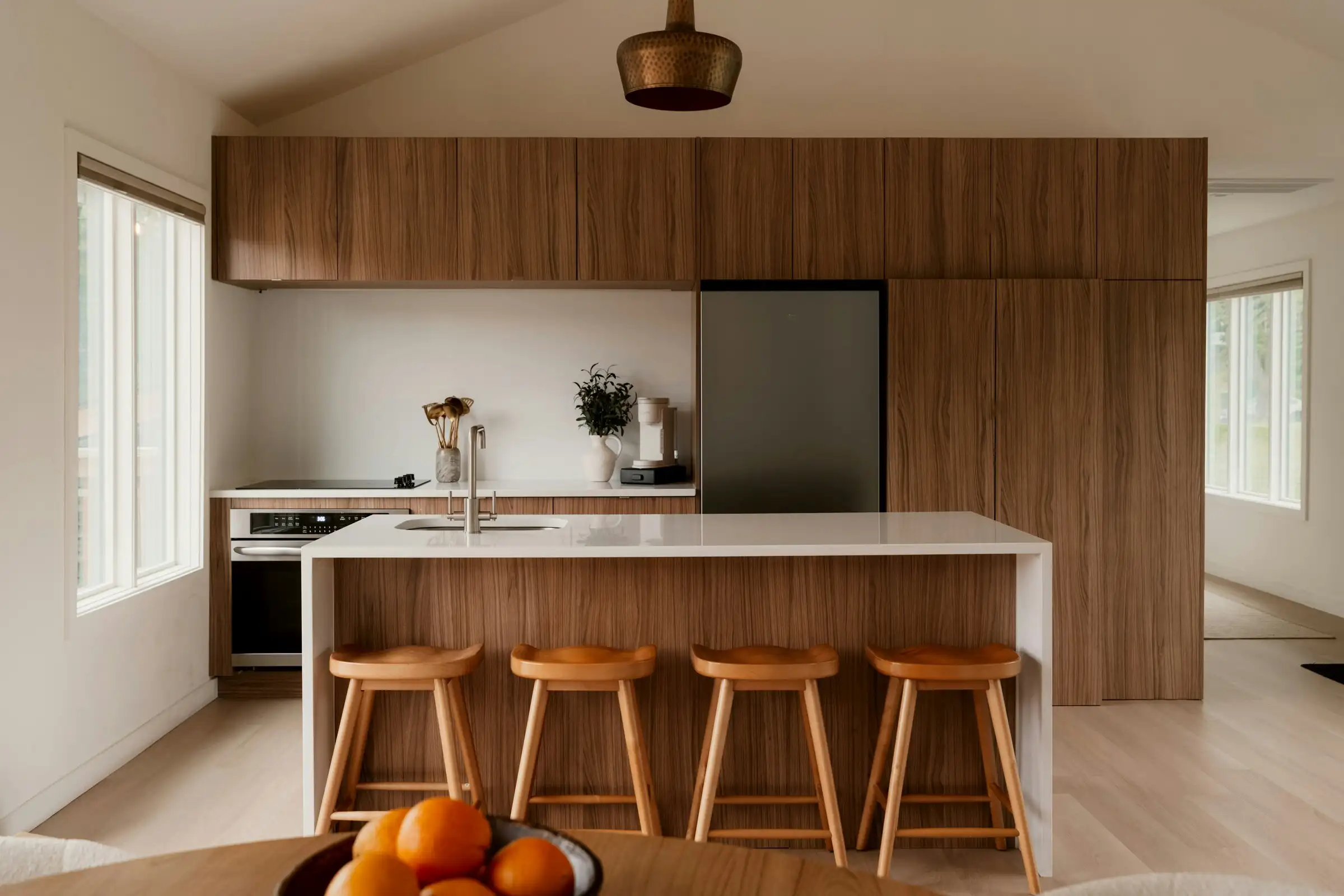 Cuisine en noyer aux façades sans poignées, îlot à plan blanc et tabourets en bois, baignée de lumière naturelle